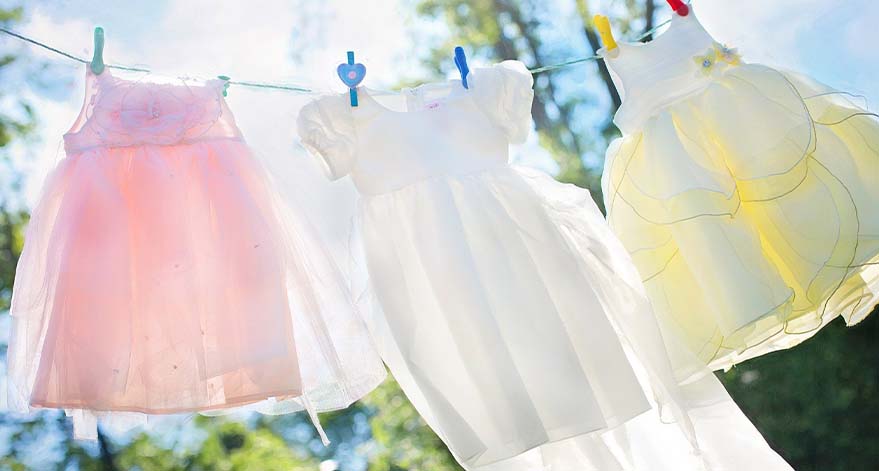 A pink dress, a white dress and a yellow dress hanging on a clothesline on a sunny day.